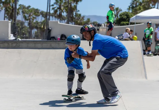 Camper riding a skateboard is helped by a counselor holding their hands