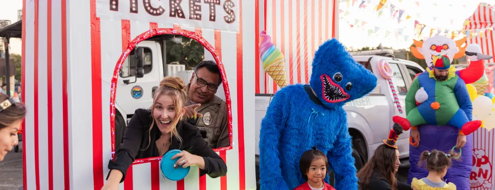 Trunk or Treat - Parks and Recreation Staff posing in a circus ticket booth and a creepy blue costume figure taking a photo with a little kid in a spiderman costume